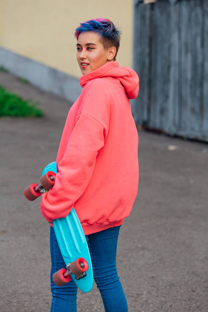 Stylish young woman with short colored hair holding blue plastic skateboard in hand. Youth concept. - Фото, изображение