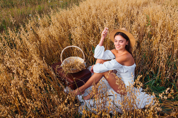 Rural Countryside Scene. Young beautiful woman with long hair dressed in white blouse and straw hat sitting at golden oat field holding basket with ears of oats. - Fotoğraf, Görsel