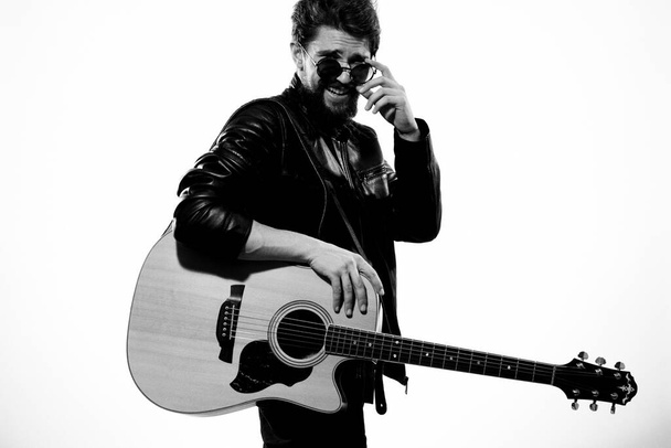 Мужчина играет на гитаре в черной кожаной куртке в солнечных очках на светлом фоне - Фото, изображение