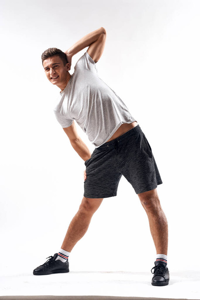A sports man in shorts and a full-length T-shirt does exercises on a light background - 写真・画像