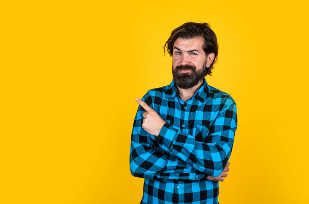 Portrait of a smiling bearded man in checkered shirt pointing fingers away at copy space over yellow background, presenting product - Valokuva, kuva