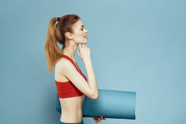 Sportieve vrouw trainingsmat in handen Rode tank top oefening blauwe achtergrond - Foto, afbeelding