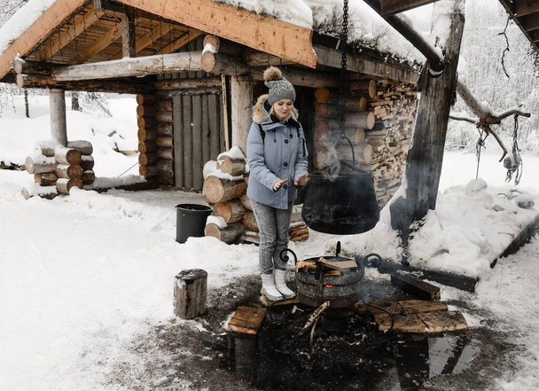 una chica con una chaqueta azul se calienta las manos sobre una hoguera donde se cocina un caldero contra una choza de madera. Karelia invierno
 - Foto, imagen