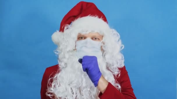 Man like a Santa Claus in medical protective mask and gloves coughs. Coronavirus COVID and Christmas, New year concept. Pandemic. Blue background - Кадри, відео