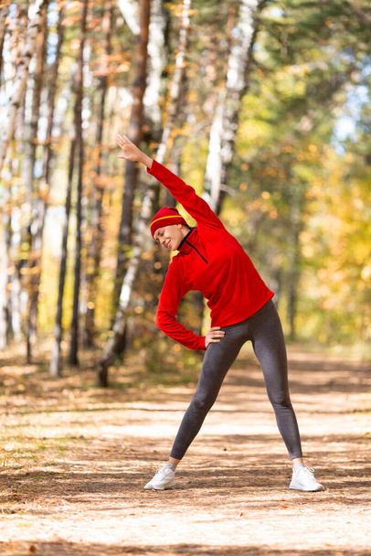 Warm up before jogging. Caucasian woman in red sportswear and a cap does side bends in the forest in autumn outdoor, vertical photo, selective focus - Fotó, kép