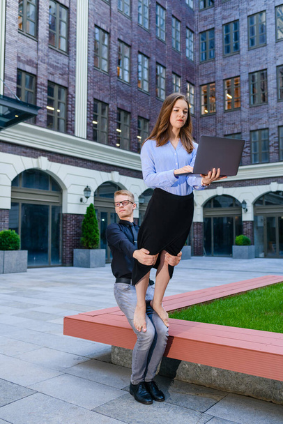 Joven hombre sostiene piernas de mujer en ropa de oficina con portátil, concepto de acroyoga para trabajar freelancer personas en el fondo del centro de negocios - Foto, imagen