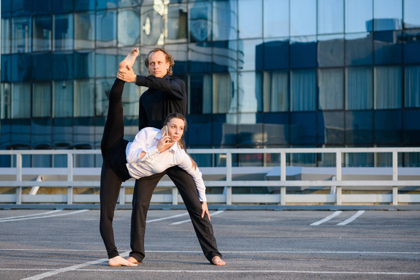 Man and woman make acroyoga element with female leg up, she talk by phone, business and sport concept at modern glass building background - Photo, Image