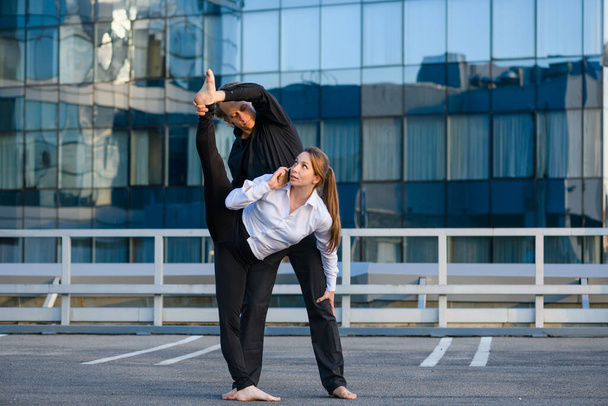 Гибкая женщина делает элемент акро-йоги, растягивая с мужчиной базовый партнер во время разговора по мобильному телефону на городском фоне, спорт и бизнес - Фото, изображение