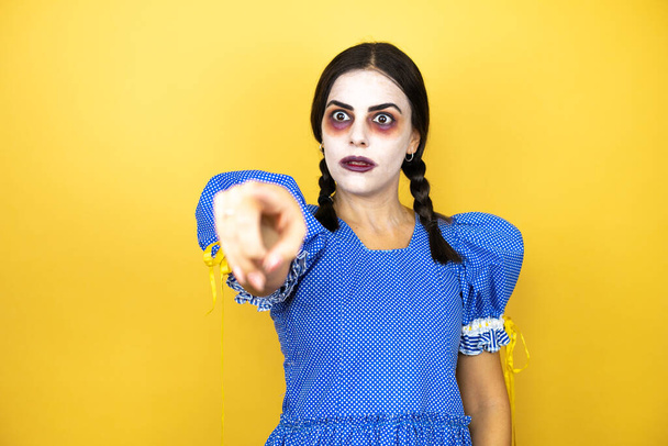 怖い人形のハロウィンの衣装を身に着けてる女が黄色い背景に指を向けて驚いて口を開けて驚いた表情正面に何か - 写真・画像