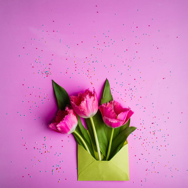 Three pink tulips in a light green envelope on a pink background decorated with sweet confetti. - Fotografie, Obrázek
