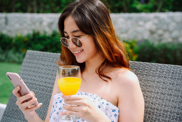 Güzel Asyalı kadın gözlük takıyor. Akıllı telefon kullanıyor ve oteldeki plaj sandalyesinde turuncu cam tutuyor.. - Fotoğraf, Görsel