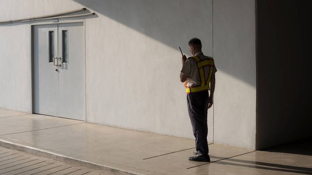 Asian security guard in safety vest walking on sidewalk and using walkie talkie or portable radio transmitter with sunlight and shadow on surface of staff room door in grey cement wall background - Photo, Image