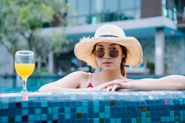 Bella donna asiatica viaggiatore indossare occhiali da sole e cappello rilassarsi in piscina con vetro di succo d'arancia. - Foto, immagini