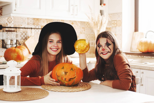 Due ragazze a casa in cucina in costume e trucco per Halloween siedono con una zucca, giocano e ridono. I bambini felici si stanno preparando per Halloween. - Foto, immagini