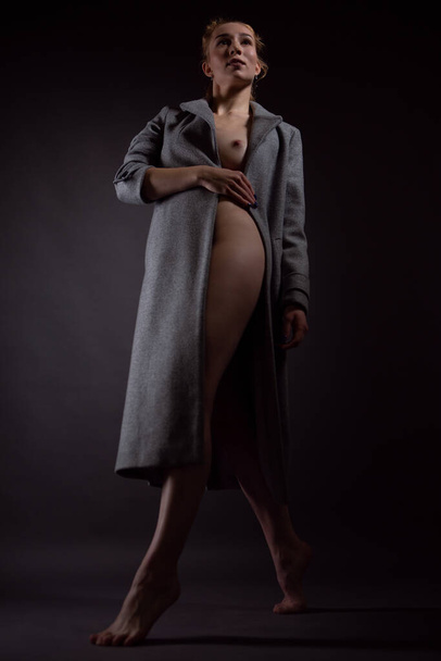 Slender naked girl in a coat poses for the camera on a dark background - Foto, afbeelding