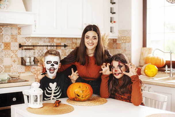 Šťastné děti slaví Halloween v kuchyni doma v kostýmech a make-up s dýňovou lucernou Jack dívá na kameru a usmívá - Fotografie, Obrázek