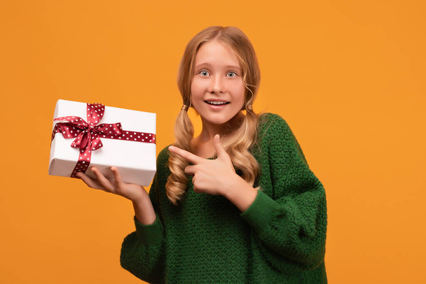 魅力的なブロンドの女の子のイメージ12-14歳暖かい緑のセーターで笑顔と赤い弓で現在のボックスを保持します。スタジオ撮影、黄色の背景。新年の女性の誕生日の休日の概念 - 写真・画像