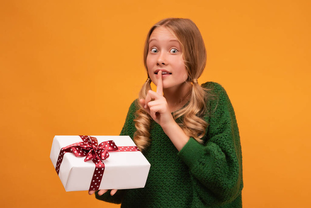Image of charming blonde girl 12-14 years old in warm green sweater holding present box with red bow. Studio shot, yellow background, isolated. New Year Women's Day birthday holiday concept - Φωτογραφία, εικόνα
