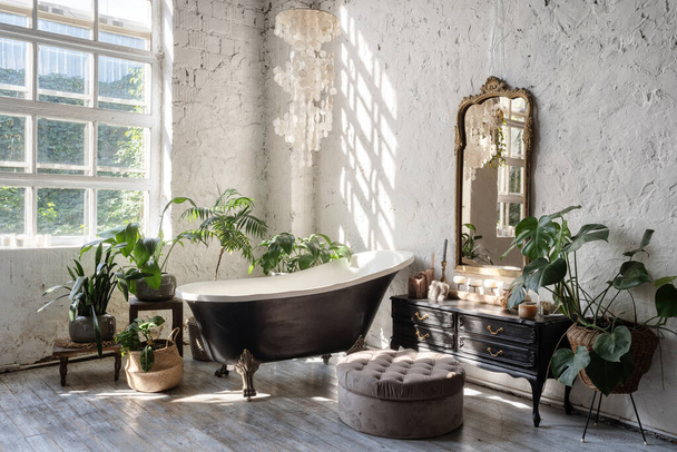 Вид збоку на білий затишний санвузол з чорною ванною, дерев'яний класичний комод, дзеркало, декор, рослини та дизайн інтер'єру в стилі бохо. Концепція комфортної і декорованої кімнати вдома
 - Фото, зображення