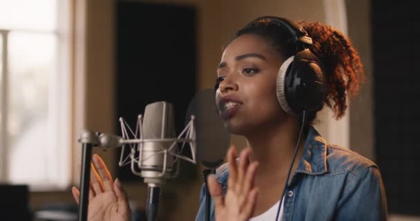 Afrikai amerikai női zenész fülhallgatóban énekel dalt mikrofonnak a hangstúdióban, lassított felvételen - Felvétel, videó