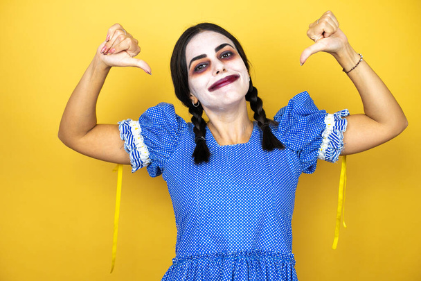 женщина в страшном костюме куклы Хэллоуина на желтом фоне выглядит уверенно с улыбкой на лице, указывая на себя пальцами гордым и счастливым. - Фото, изображение