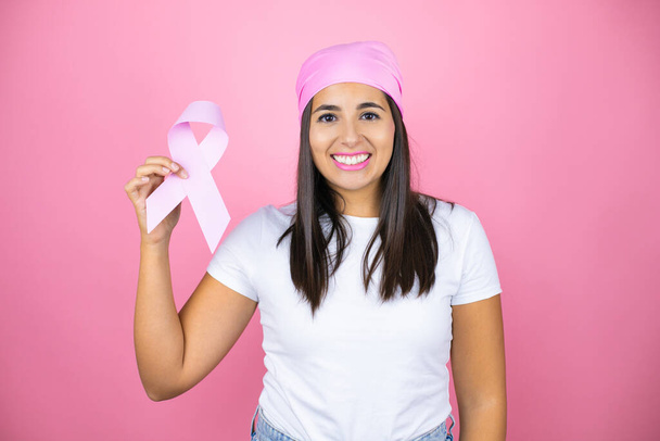 Νέα όμορφη γυναίκα φορώντας ροζ μαντίλα κρατώντας κορδέλα για τον καρκίνο Brest πάνω από απομονωμένο ροζ φόντο με ένα χαρούμενο πρόσωπο στέκεται και χαμογελά με αυτοπεποίθηση χαμόγελο που δείχνει τα δόντια - Φωτογραφία, εικόνα