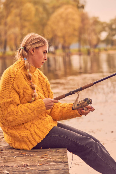 A girl with pigtails in a bright yellow knitted sweater sits on a wooden bridge over the lake and catches fish. - Foto, Bild