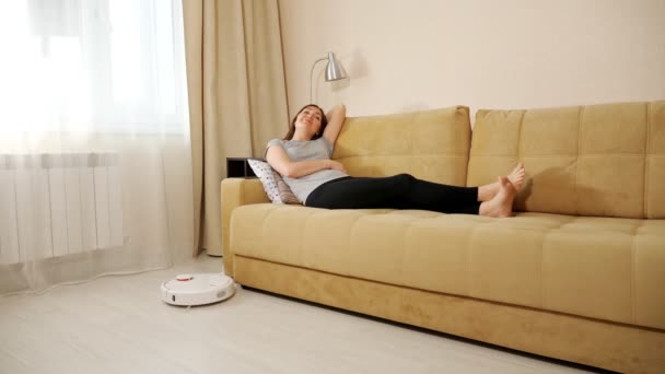 Glückliche Frau liegt auf Sofa, während Saugroboter Staubsauger Boden putzt - Filmmaterial, Video