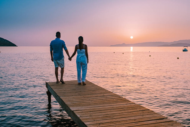 Kreta Grecja, młoda zakochana romantyczna para siedzi i przytula się na drewnianym molo na plaży o wschodzie słońca ze złotym niebem. Koncepcja wakacji i podróży. Romantyczna młoda para randkowa nad morzem - Zdjęcie, obraz