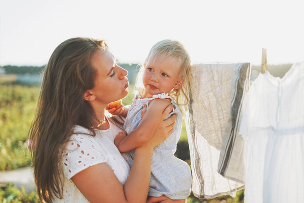 Mladá žena maminka s dítětem dívka na ruce objetí na dvorku chalupy v západu slunce - Fotografie, Obrázek