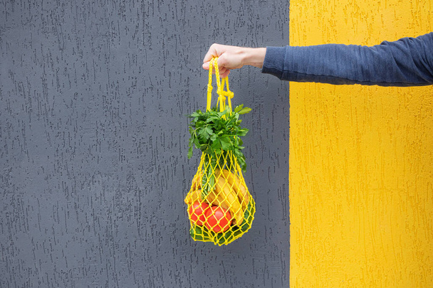 Sárga húros táska uborkával, paradicsommal, banánnal és gyógynövényekkel a kezében. Fényes fénykép piros, sárga, zöld tónusokkal. Fenntarthatóság, nulla hulladék, műanyagmentes koncepció, vegetarianizmus, egészséges élelmiszer. - Fotó, kép