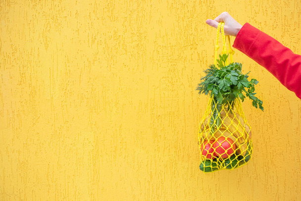キュウリ、トマト、バナナ、ハーブが入った黄色のストリングバッグ。赤、黄色、緑のトーンで明るい写真。持続可能性、廃棄物ゼロ、プラスチックフリーコンセプト、菜食主義、健康食品. - 写真・画像