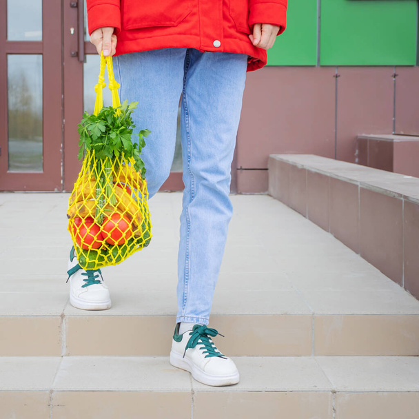 Желтая струнная сумка с огурцами, помидорами, бананами и травами в руках девушки в синих джинсах, красная куртка. Яркое фото в красных, желтых и зеленых тонах. Устойчивость, нулевые отходы, пластиковая свободная концепция. - Фото, изображение