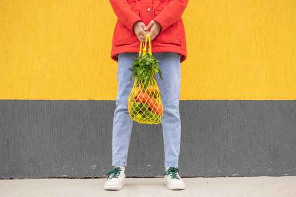 Sárga húrtáska uborkával, paradicsommal, banánnal és gyógynövényekkel egy kék farmeres lány kezében, piros kabátban. Fényes fénykép piros, sárga és zöld színekben. Fenntarthatóság, nulla hulladék, műanyagmentes koncepció. - Fotó, kép
