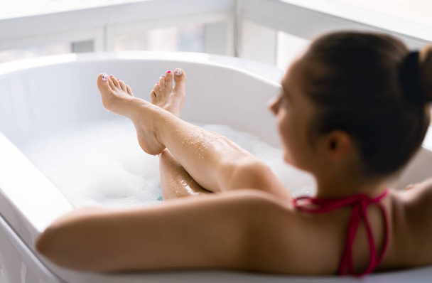 Kobieta w kąpieli z bąbelkami po pedicure i lakierowaniu paznokci u stóp. Kobieta relaksująca się w wannie. Czyste mokre nogi i stopy w wannie z pianką. Rozpieszczanie i leczenie w spa. Dziewczyna korzystających z ciepłej wody rano - Zdjęcie, obraz