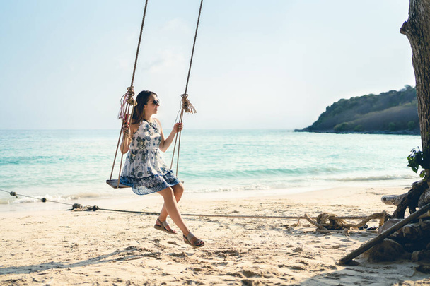 Swing σε τροπικό παράδεισο παραλία. Γυναίκα απολαμβάνοντας τη ζωή και τις καλοκαιρινές διακοπές σε ηλιόλουστο νησί στην Ταϊλάνδη. Όμορφη νεαρή κοπέλα με φόρεμα διασκεδάζει. Χαλαρωτικά κύματα του ωκεανού και άμμος. Ξέγνοιαστες διακοπές τρόπο ζωής. - Φωτογραφία, εικόνα