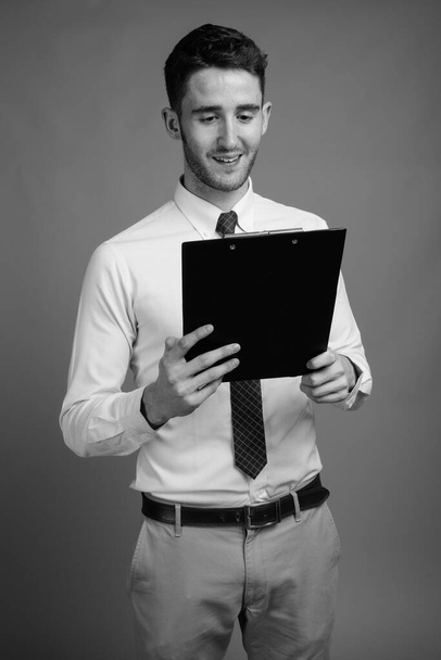 Studioaufnahme eines jungen, gut aussehenden Geschäftsmannes mit Klemmbrett vor grauem Hintergrund in Schwarz-Weiß - Foto, Bild