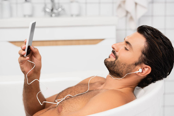 Young man in earphones taking bath and using smartphone  - Фото, изображение