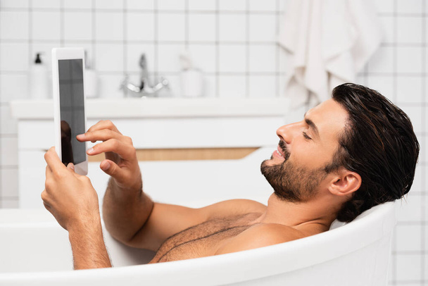 Smiling man using digital tablet with blank screen while taking bath at home  - Фото, изображение