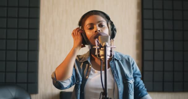 Hivatásos énekes. Fiatal fekete hölgy fülhallgatóban énekel mikrofonnak a hangstúdióban - Felvétel, videó