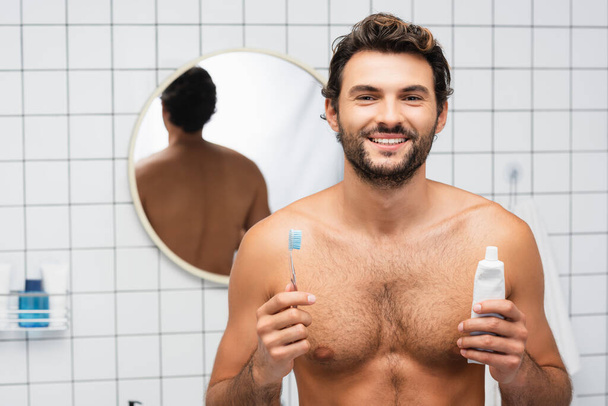 Hombre alegre sin camisa mirando a la cámara mientras sostiene la pasta de dientes y el cepillo de dientes en el baño  - Foto, Imagen