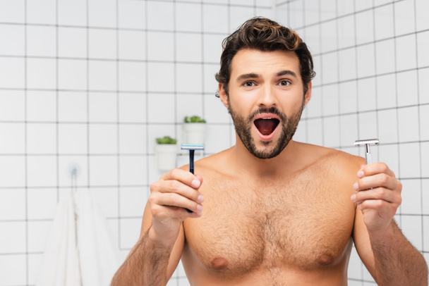 Homme torse nu excité regardant la caméra tout en tenant des rasoirs dans la salle de bain  - Photo, image