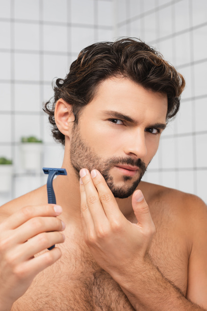 Bärtiger Mann hält Rasiermesser in der Hand und blickt in Badezimmer in die Kamera  - Foto, Bild