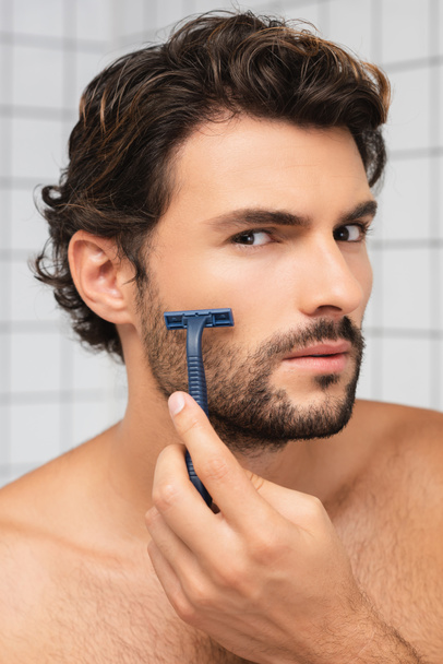 Shirtless man looking at camera while shaving in bathroom  - Valokuva, kuva