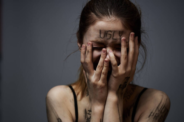 Разочарованная женщина с оскорбительными надписями на теле и в черной футболке на сером фоне  - Фото, изображение