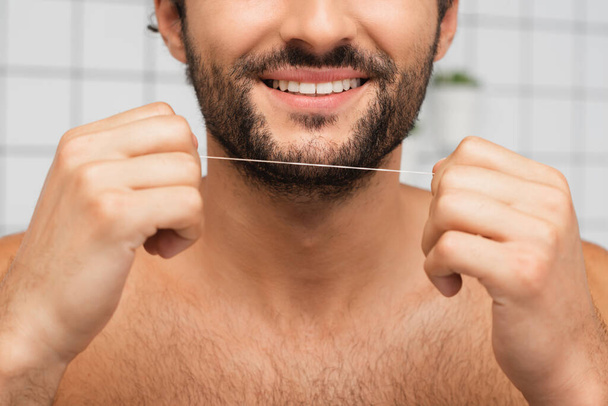 Vista recortada del hombre barbudo sonriendo mientras sostiene el hilo dental en el baño  - Foto, imagen