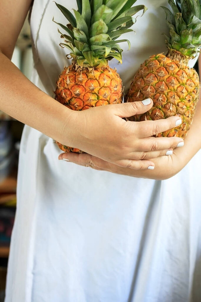 beschnittene junge Frau in weißer Kochschürze hält in ihren Händen zwei reife und saftige Ananas. - Foto, Bild