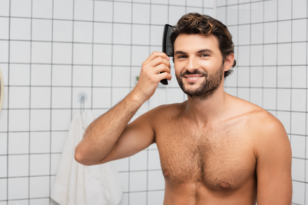 Smiling muscular man combing hair in bathroom  - Fotó, kép