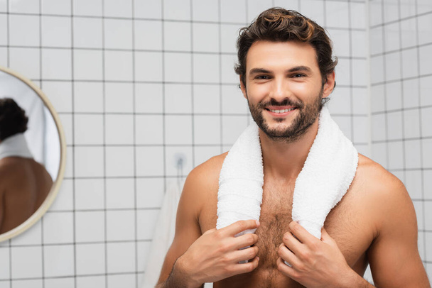 Smiling shirtless man touching towel and looking at camera in bathroom  - Φωτογραφία, εικόνα