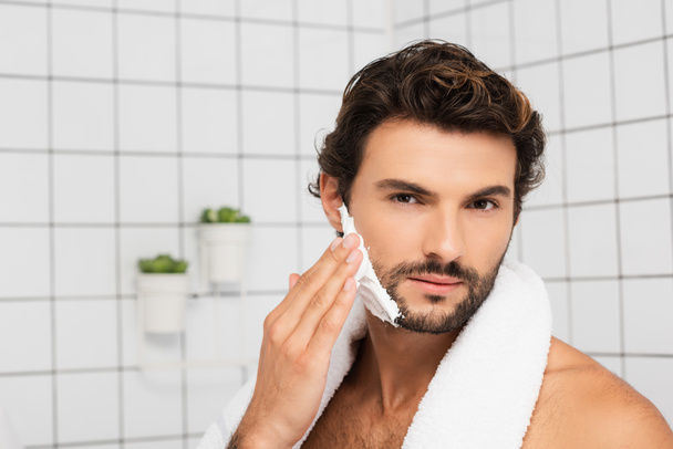 Bearded man with towel applying shaving foam in bathroom  - Фото, изображение